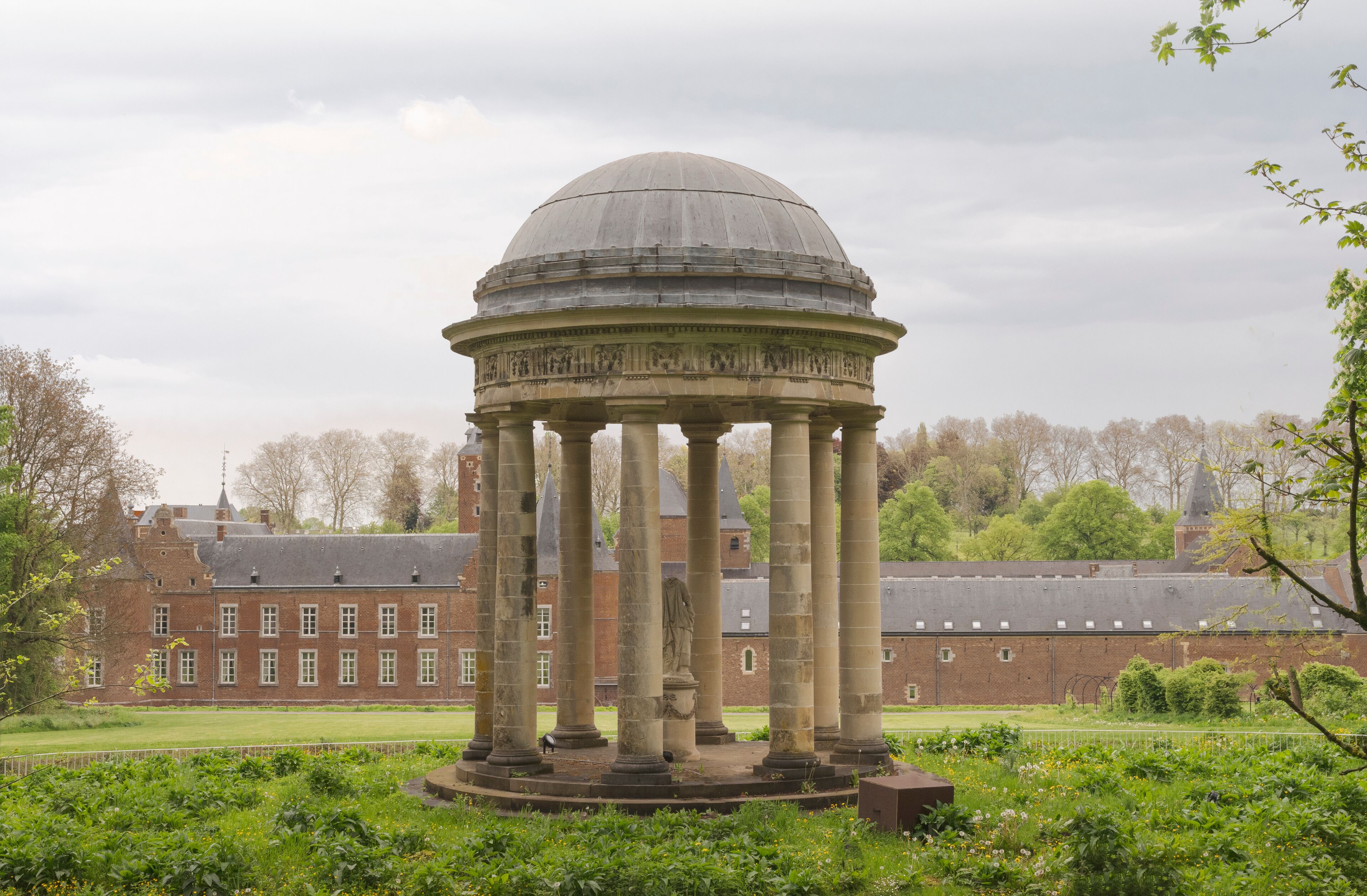 A classical pavilion with a dome and columns, surrounded by lush greenery and a large brick building in the background.