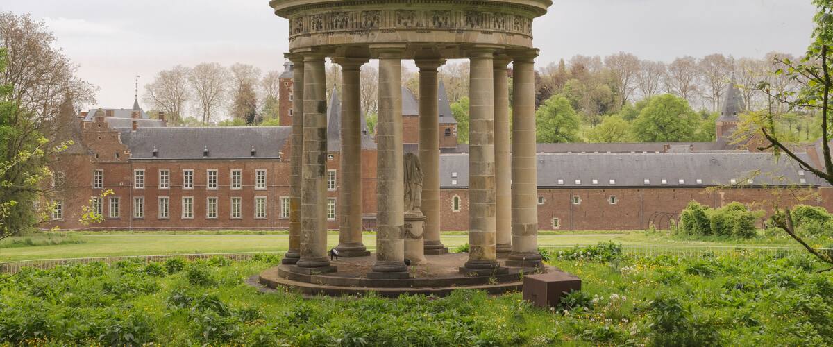 A classical pavilion with a dome and columns, surrounded by lush greenery and a large brick building in the background.
