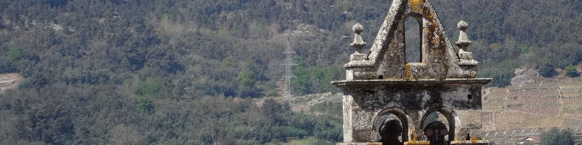 Ein steinerner Glockenturm mit Kreuz, umgeben von einem Dorf und Bergen im Hintergrund.