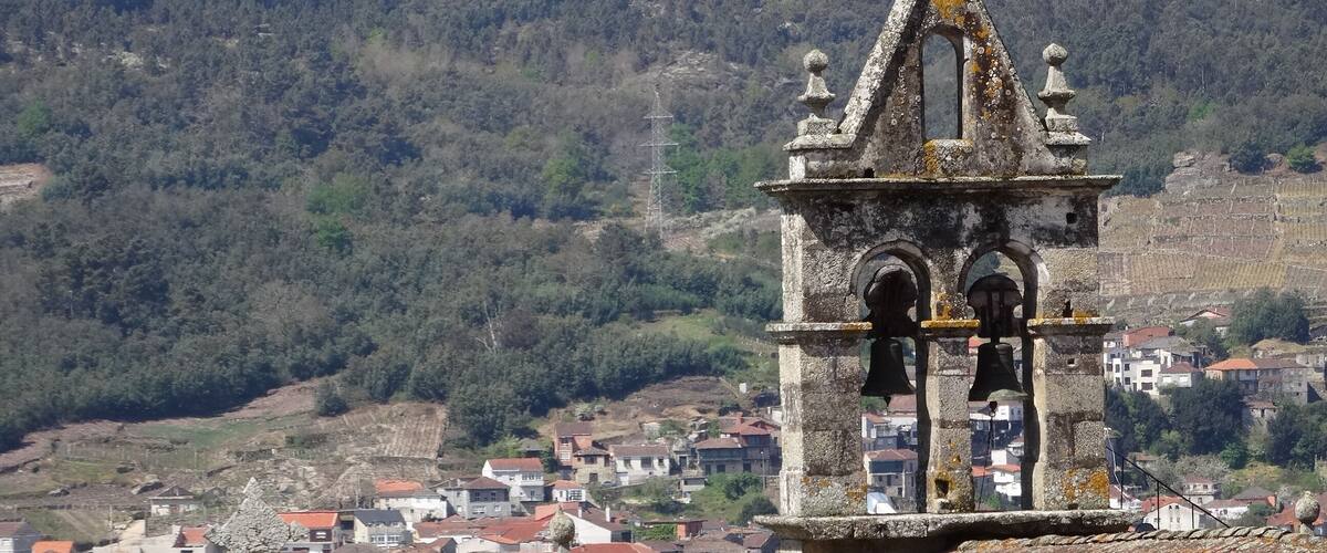 Un campanario de piedra con una cruz en la parte superior, rodeado por un pueblo y montañas al fondo.