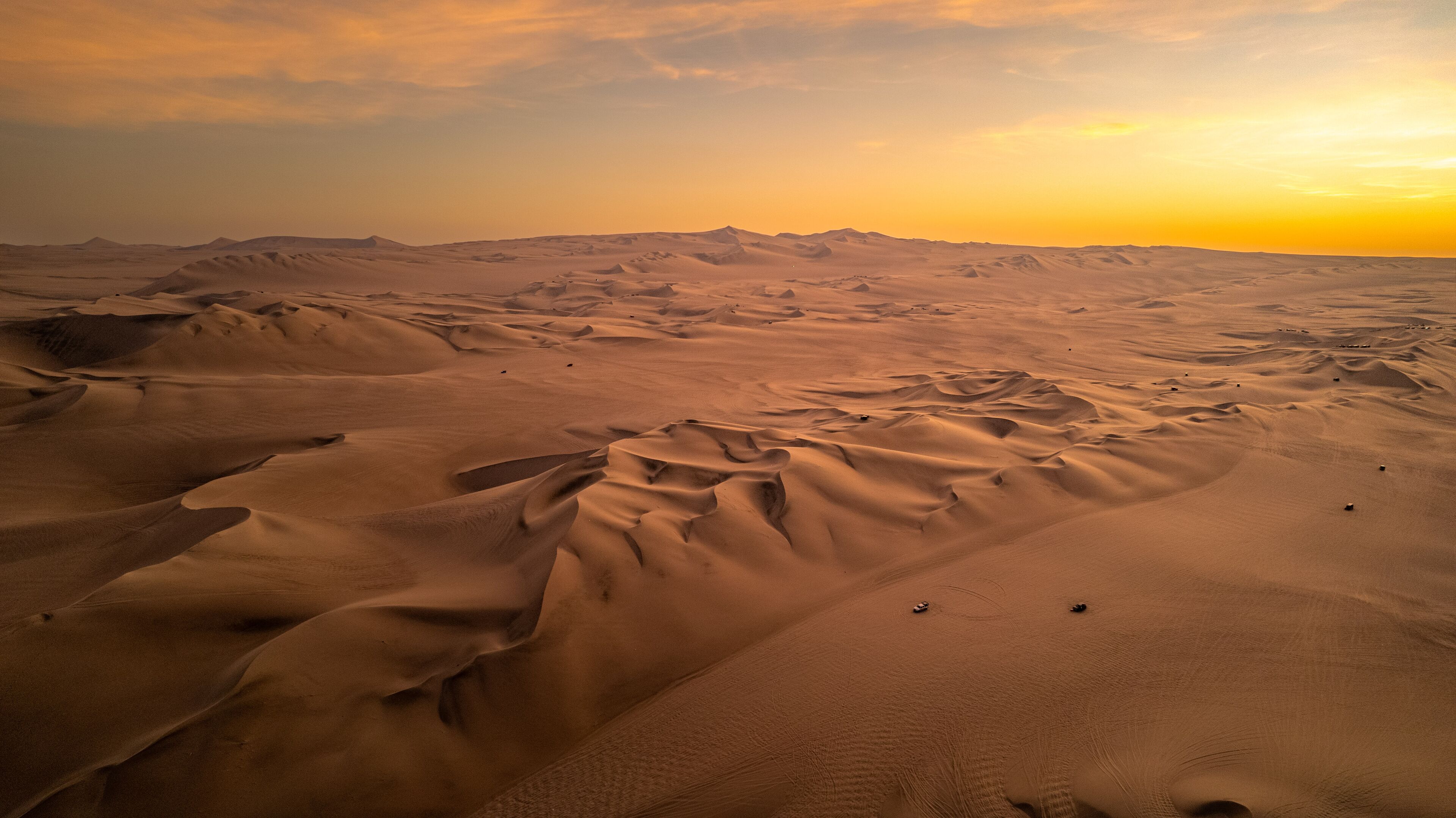 A vast desert landscape with undulating sand dunes and a setting sun.