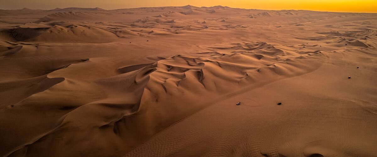 A vast desert landscape with undulating sand dunes and a setting sun.