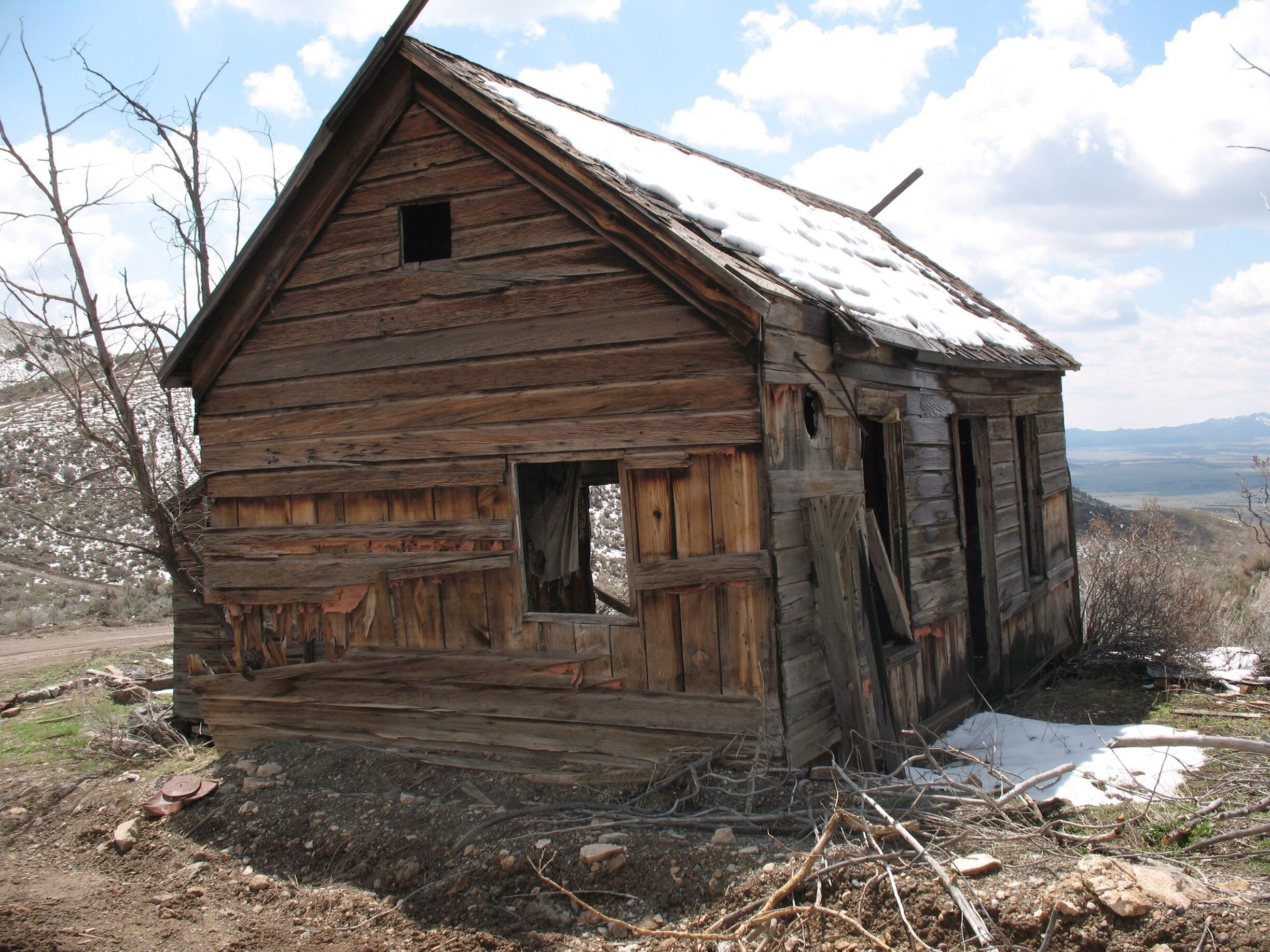 A dilapidated wooden cabin with a steep roof, surrounded by a barren landscape with sparse vegetation and a distant mountain range.