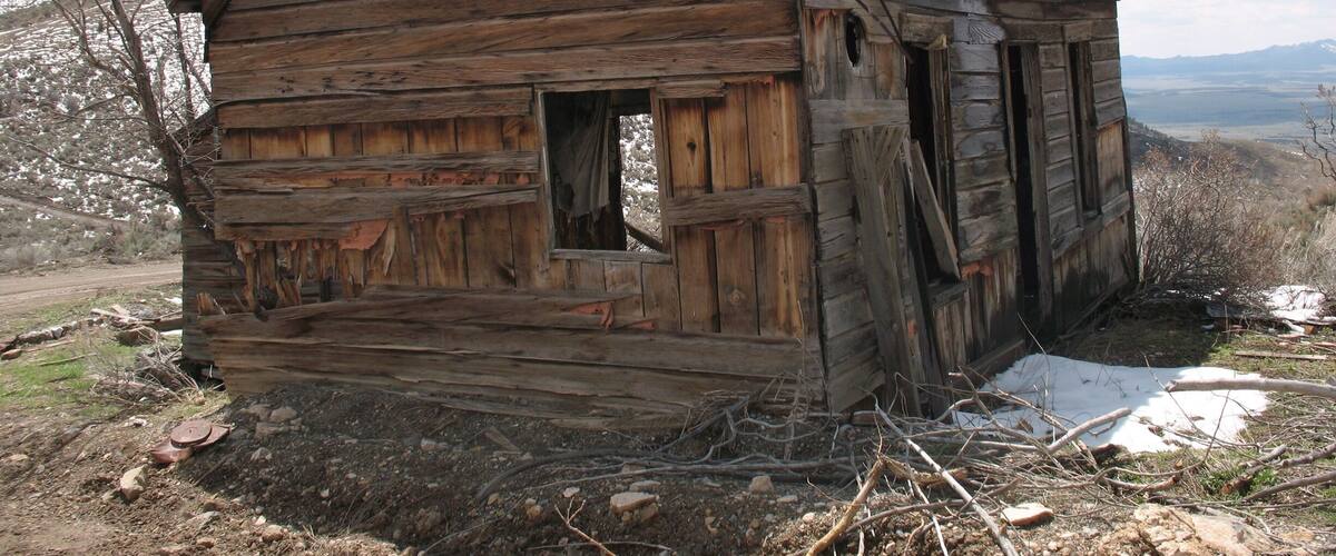 A dilapidated wooden cabin with a steep roof, surrounded by a barren landscape with sparse vegetation and a distant mountain range.
