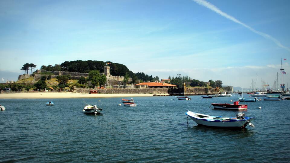 Un pueblo costero con barcos en el agua, una playa de arena y una colina con una estructura similar a un castillo.