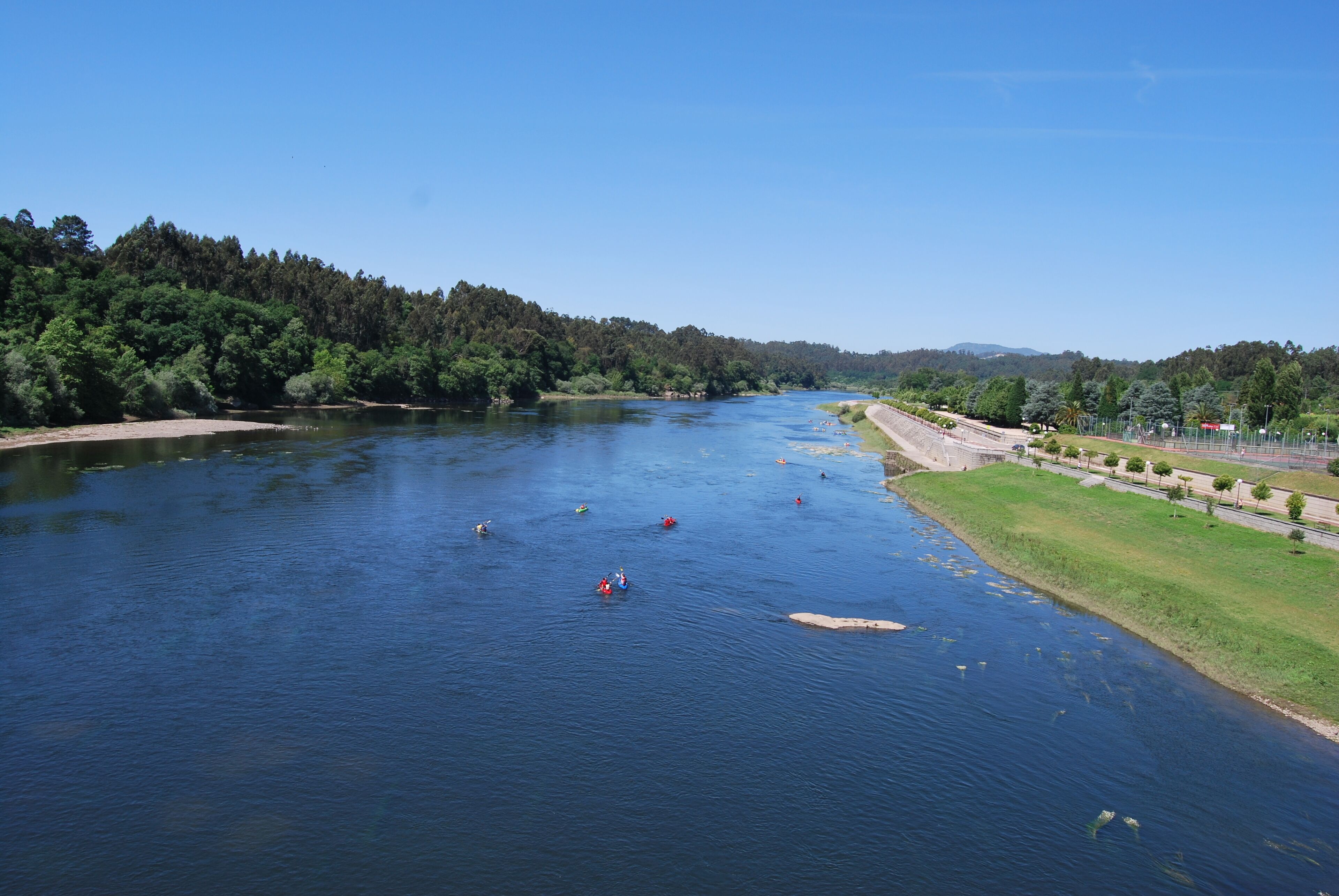 A river with kayaks, surrounded by dense forest and a paved path.