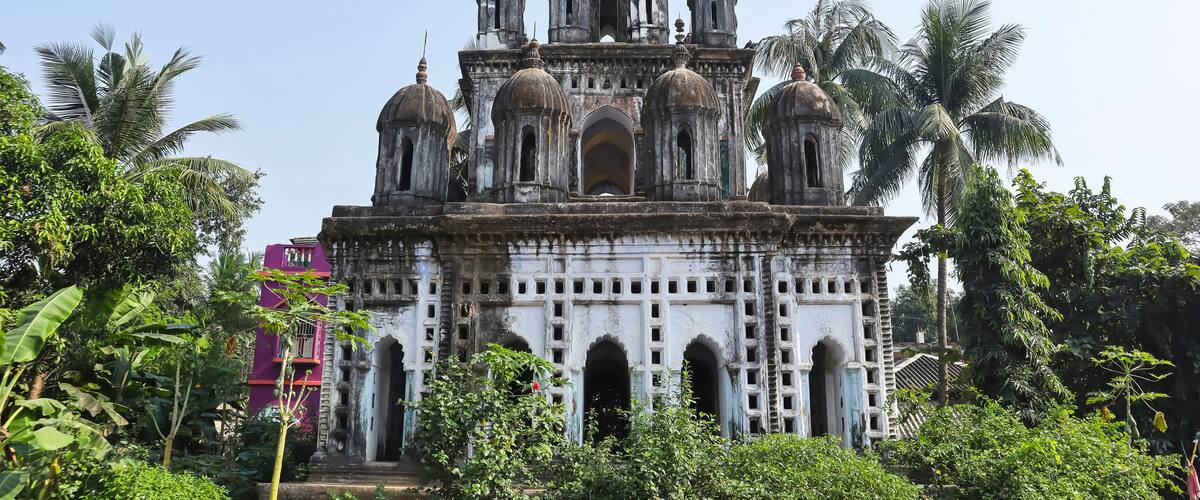 A historic temple with multiple domes and arches, surrounded by lush greenery and palm trees.
