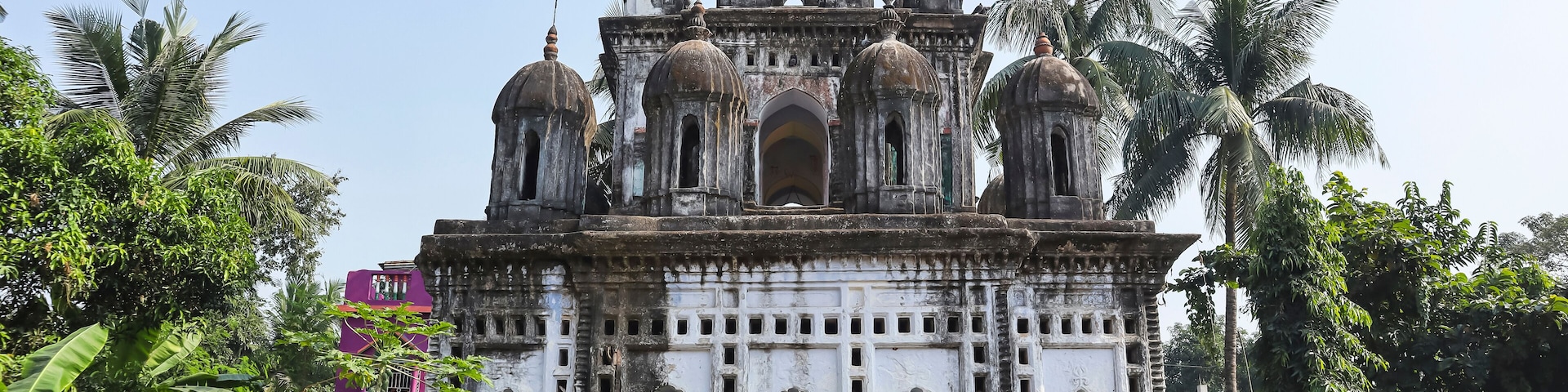 A historic temple with multiple domes and arches, surrounded by lush greenery and palm trees.