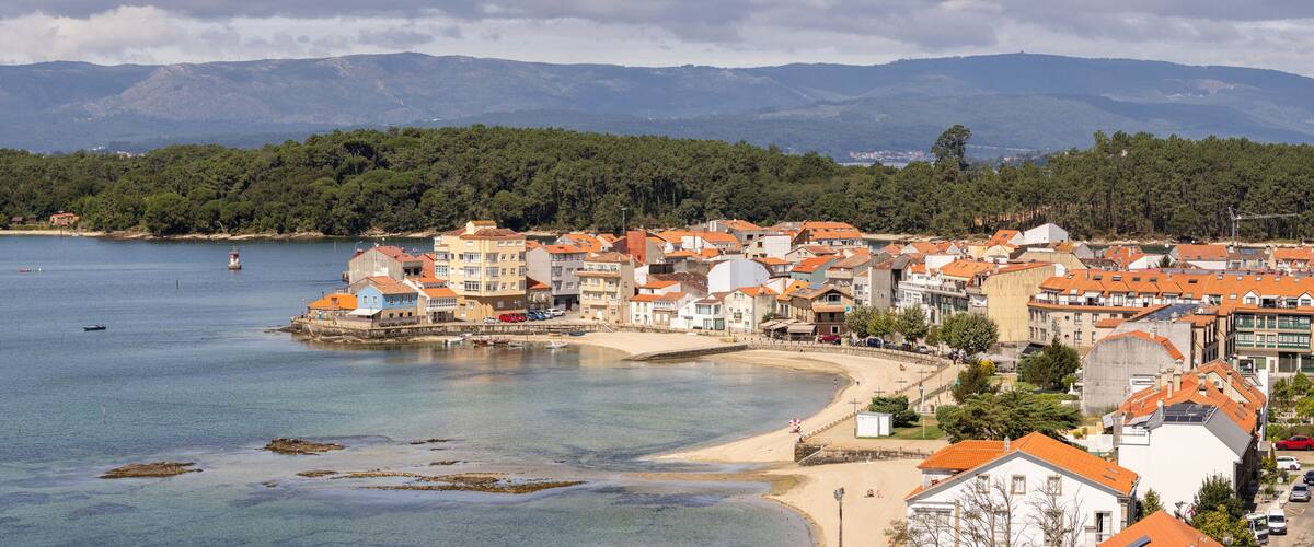 A coastal town with buildings along the shore, a sandy beach, and a forested hill in the background.