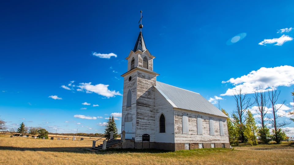 A white church with a steeple and a cross on top, surrounded by a grassy field and trees.