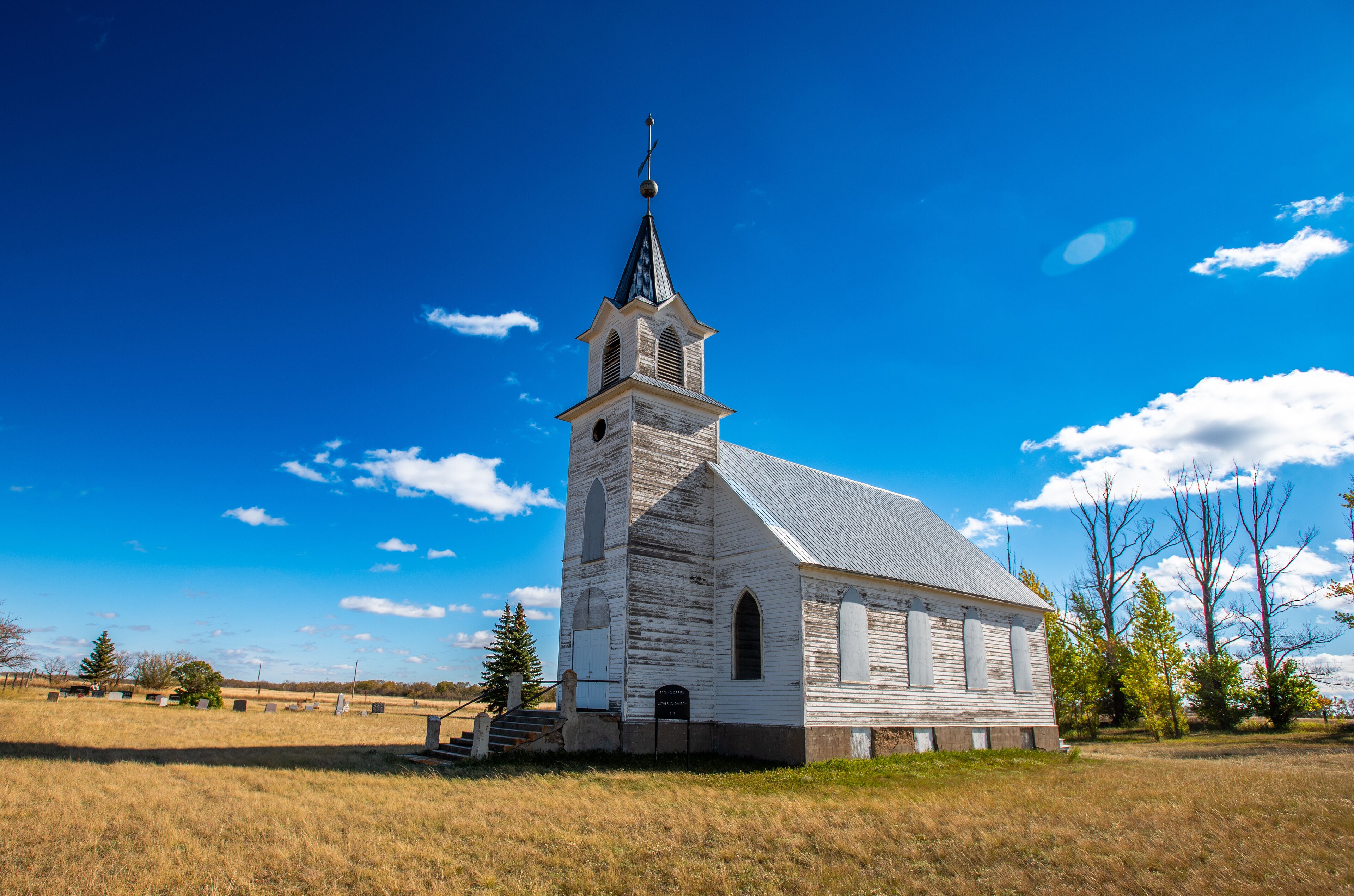 A white church with a steeple and a cross on top, surrounded by a grassy field and trees.