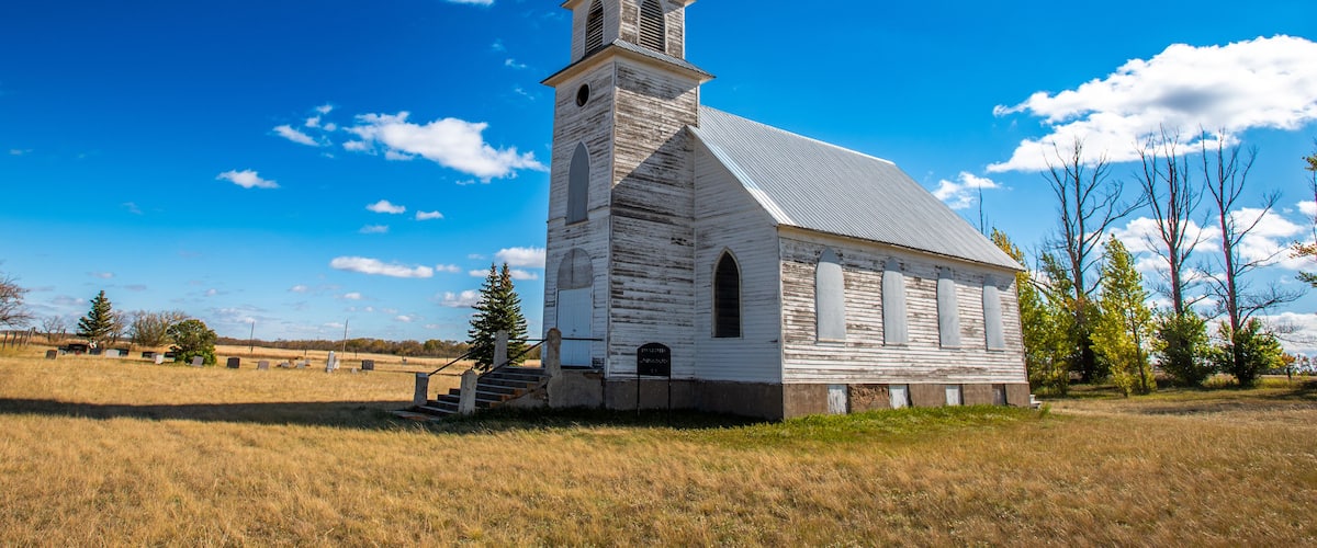 A white church with a steeple and a cross on top, surrounded by a grassy field and trees.