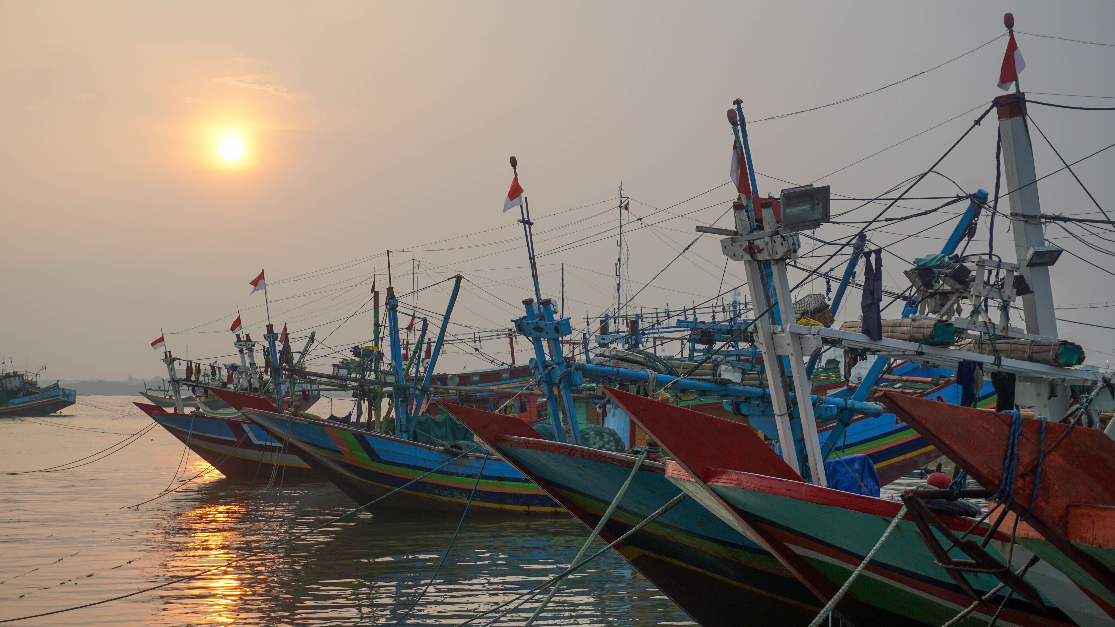 A row of colorful fishing boats docked at a harbor with the sun setting in the background.