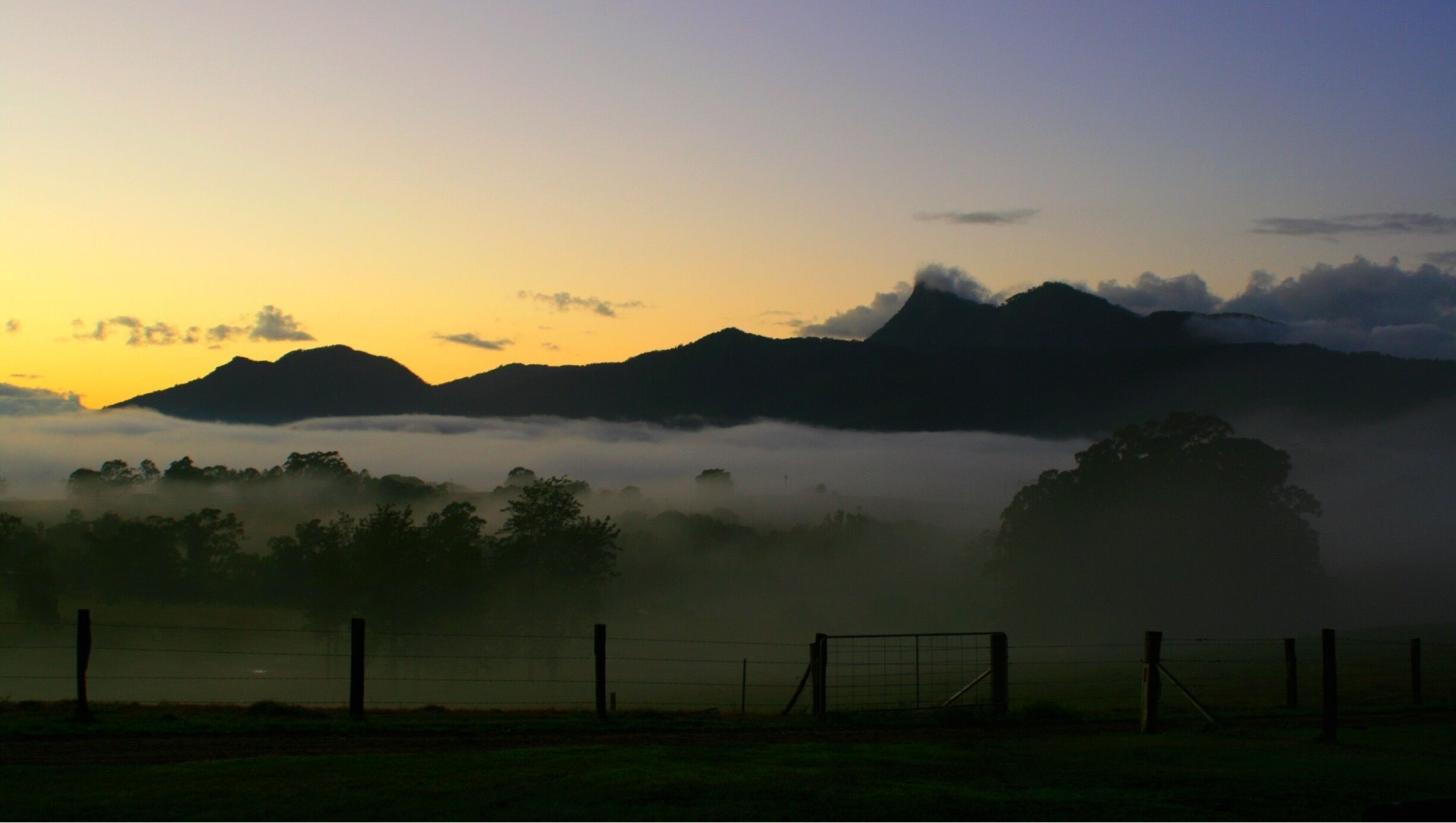 A sunrise over a mountainous landscape with a foggy valley and a fence in the foreground.