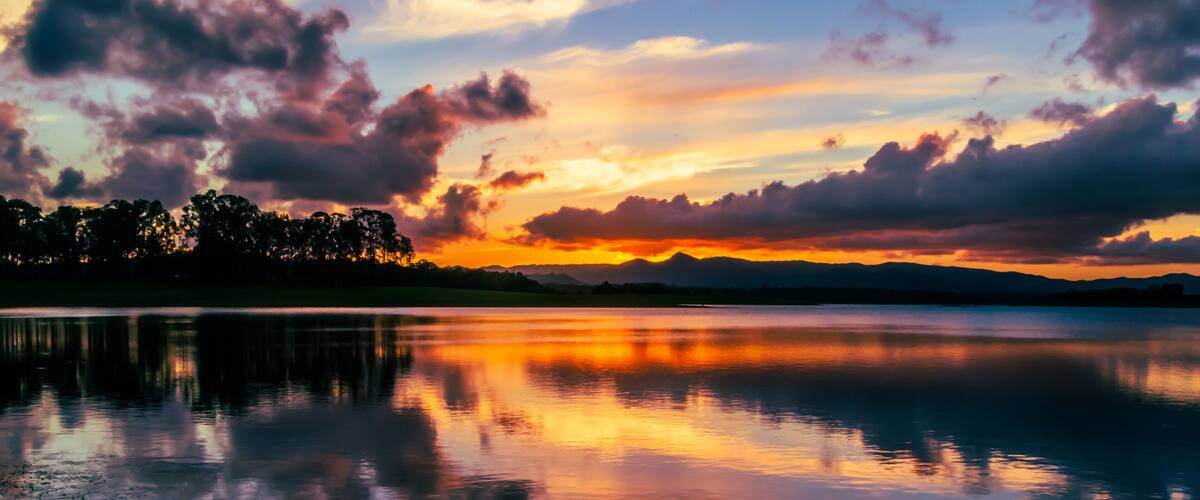 A sunset over a calm lake with mountains in the distance and a forested area along the water's edge.