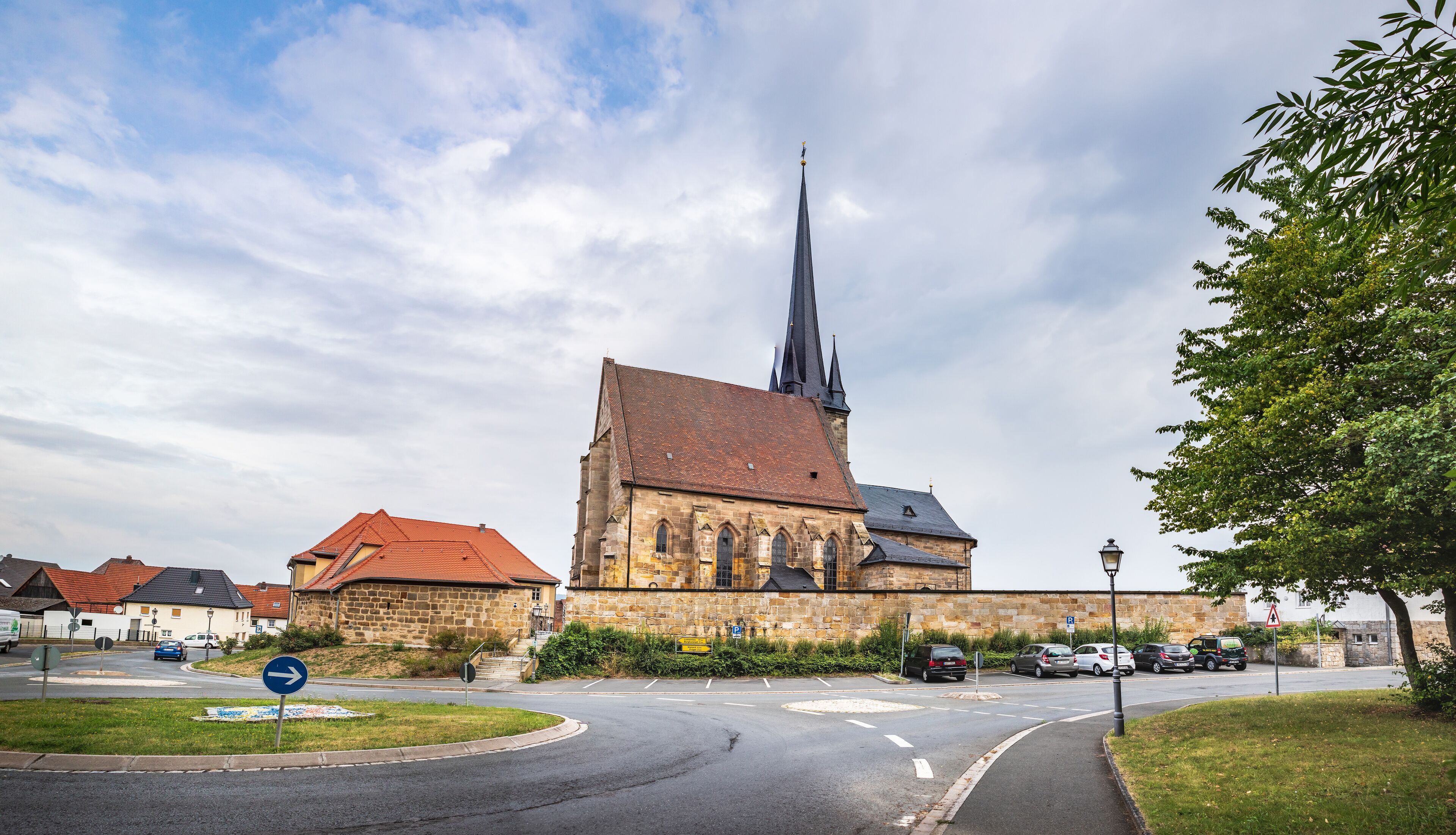 A church with a tall spire, a stone building, and a parking area with cars.