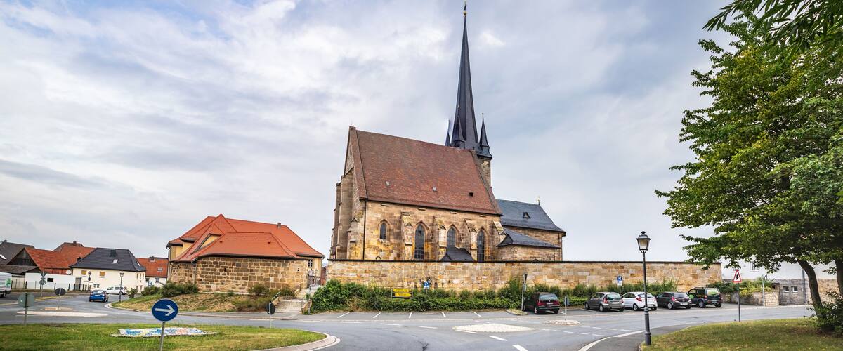 A church with a tall spire, a stone building, and a parking area with cars.