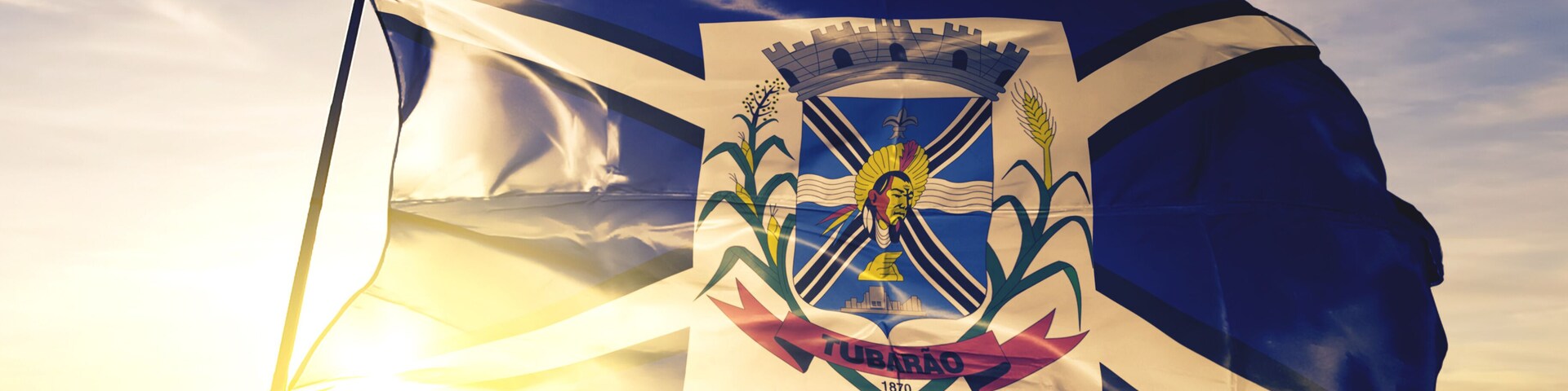 Uma bandeira com brasão, um sol e a palavra 'TUBARAO' ao fundo de um céu ao nascer do sol.