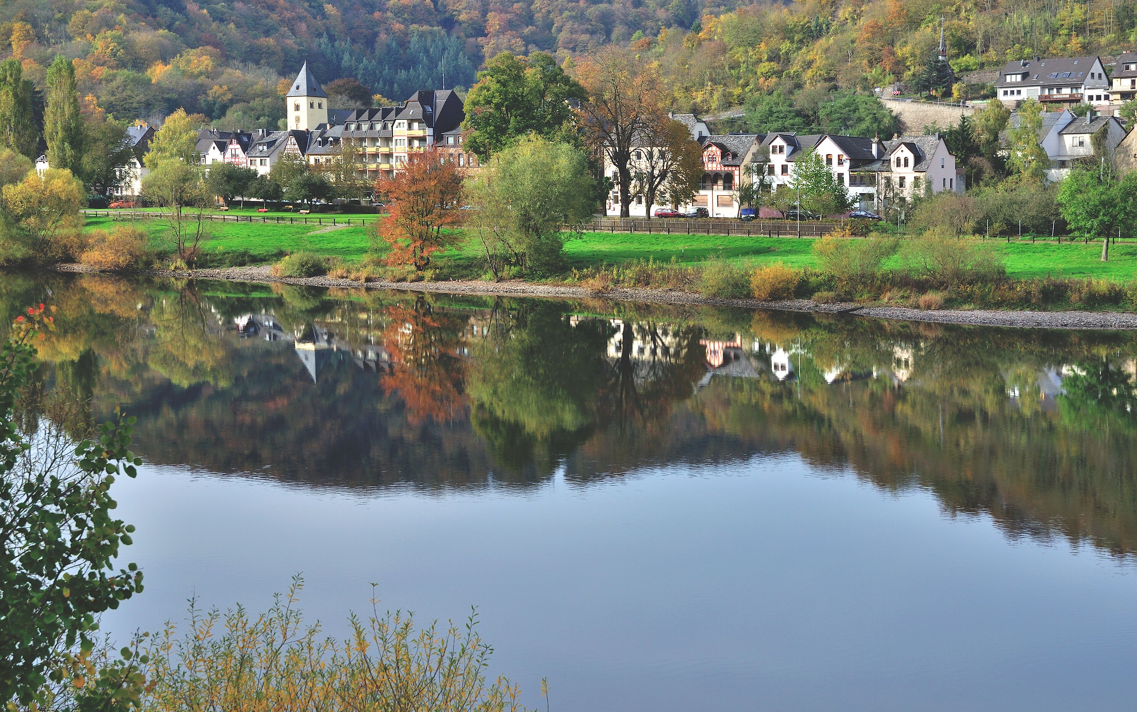 A calm lake reflecting houses and trees, surrounded by a lush green landscape and hills.