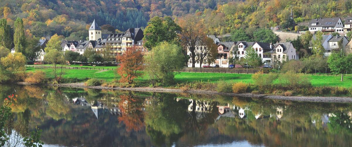 A calm lake reflecting houses and trees, surrounded by a lush green landscape and hills.
