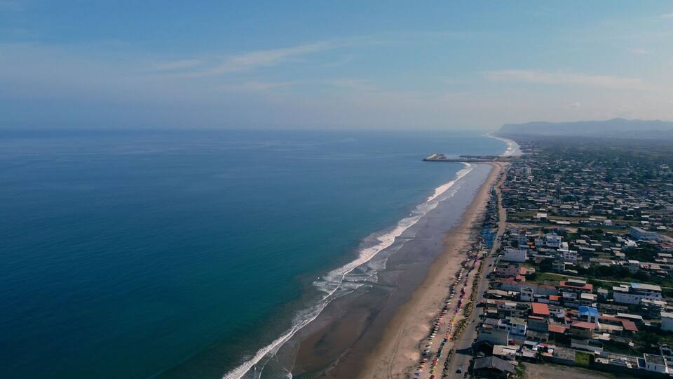 An aerial view of a coastal city with a long beach, dense housing, and a distant peninsula.