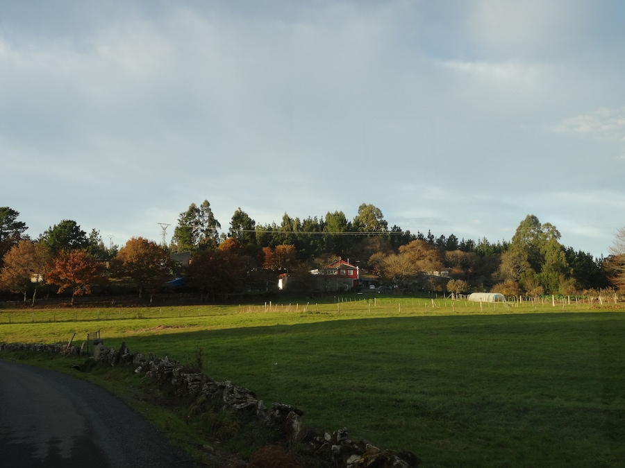 Een landelijk landschap met een stenen muur, een weg, en een heuvel met bomen en een huis.