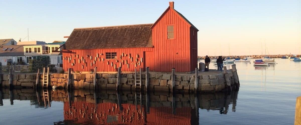 A red wooden boathouse by a calm water body with boats in the background.