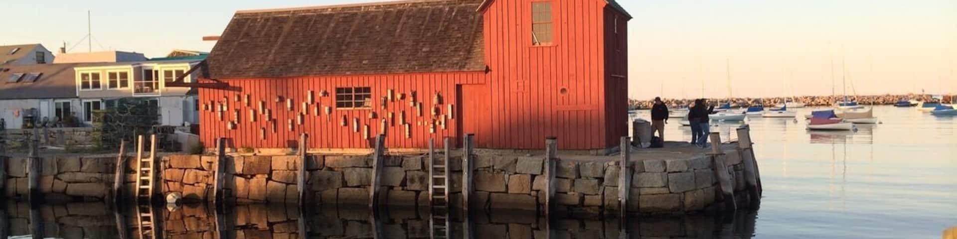 A red wooden boathouse by a calm water body with boats in the background.