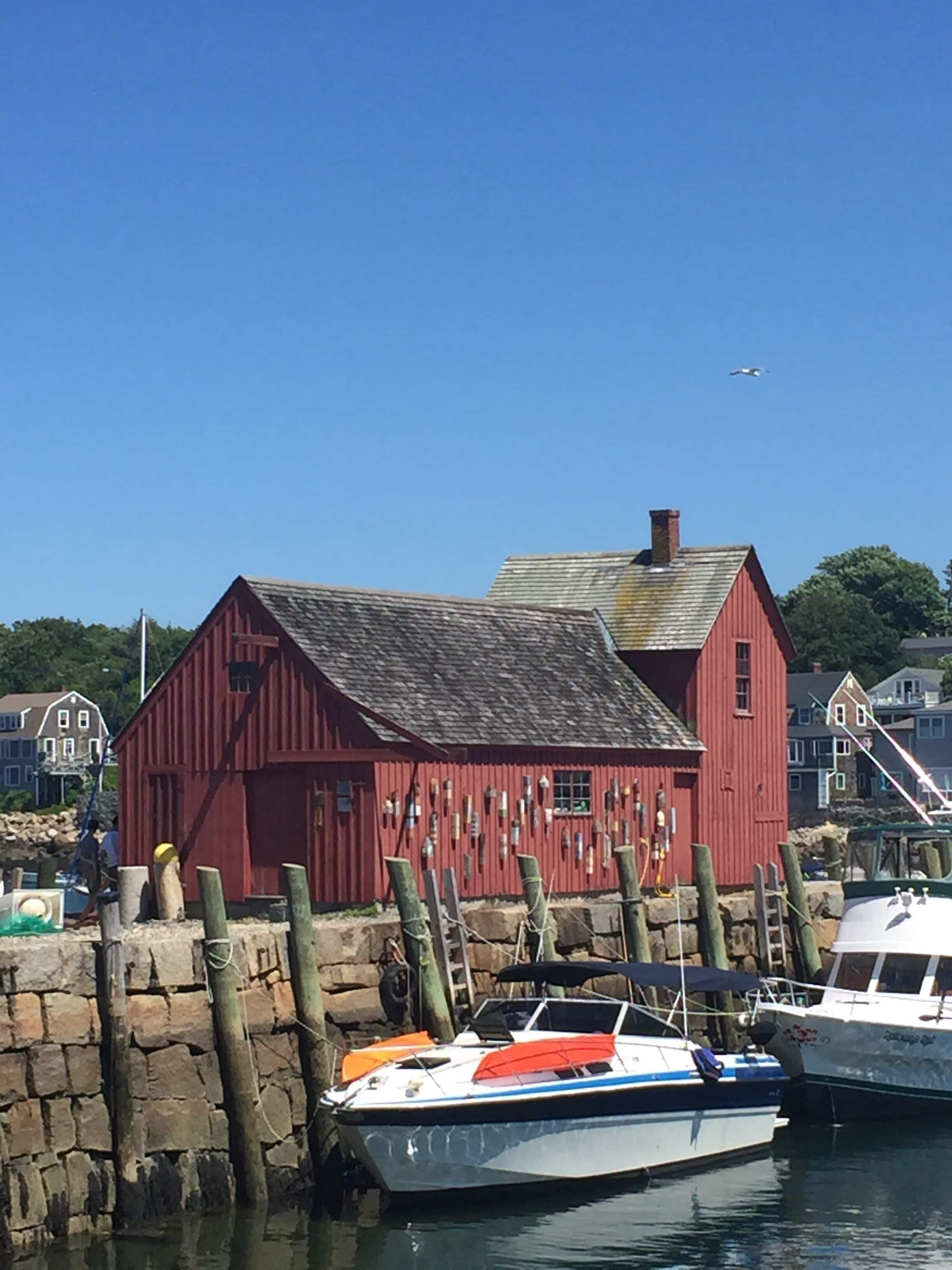 Un bâtiment rouge avec un toit en pente, un quai où sont amarrés des bateaux et un ciel bleu clair.