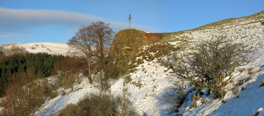 A snow-covered hill with a wooden cross on top, surrounded by trees and a clear sky.