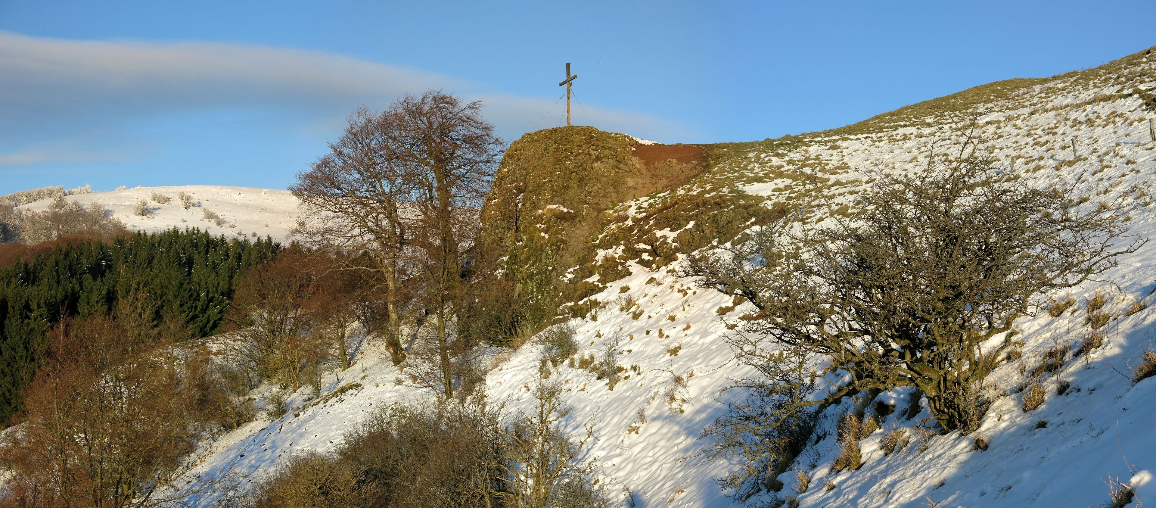 A snow-covered hill with a wooden cross on top, surrounded by trees and a clear sky.