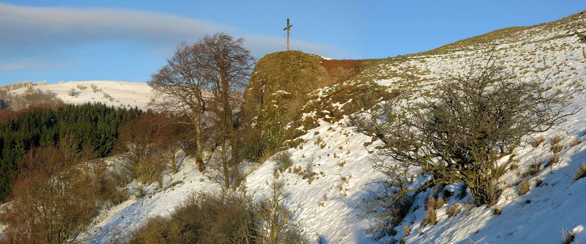 A snow-covered hill with a wooden cross on top, surrounded by trees and a clear sky.