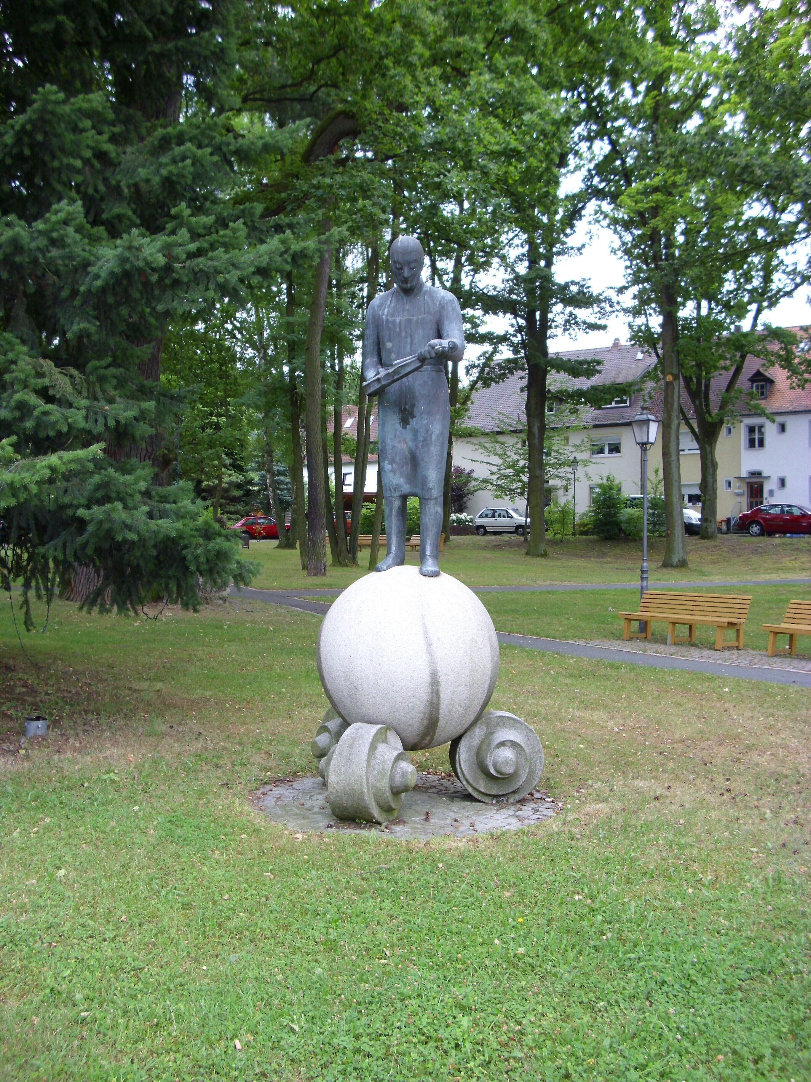 A statue of a man holding a tool, surrounded by trees and benches in a park.
