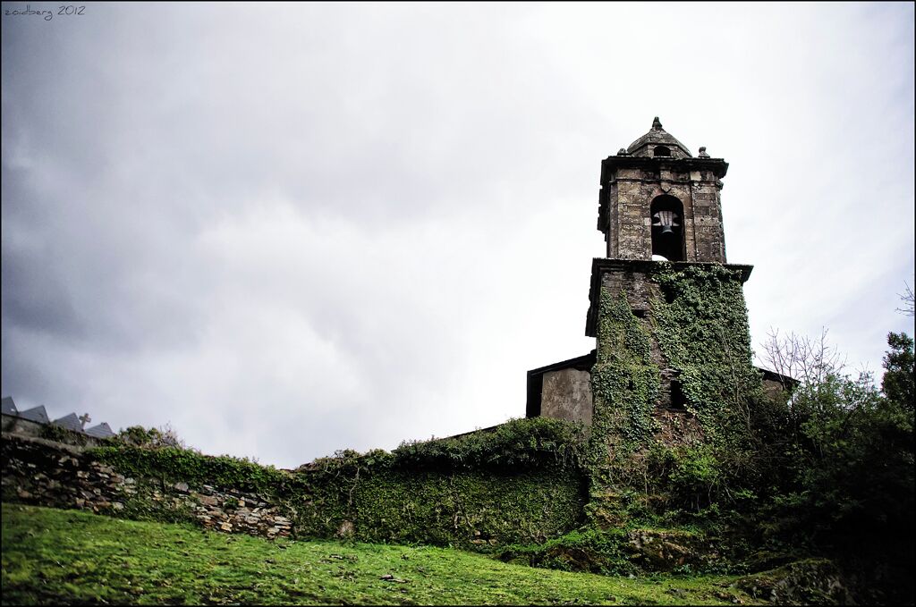 A church tower with a clock, surrounded by greenery and a cloudy sky.