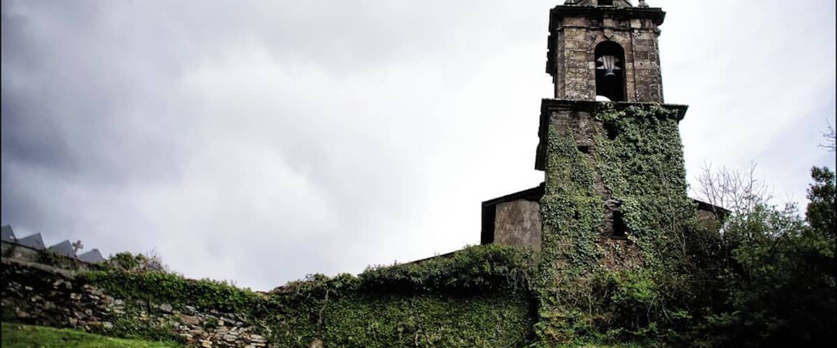A church tower with a clock, surrounded by greenery and a cloudy sky.