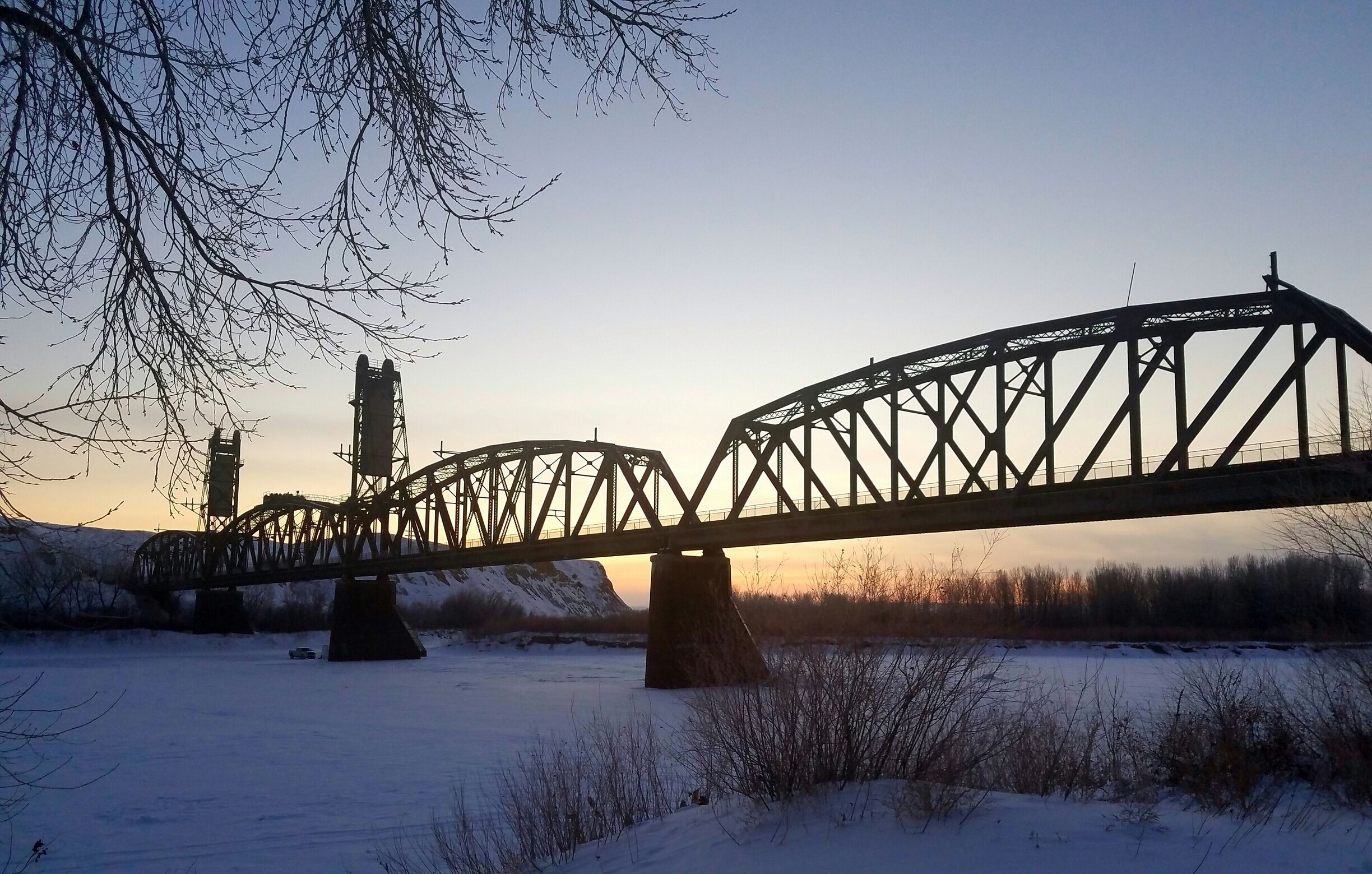 A steel bridge over a snow-covered river with bare trees and a distant hill.