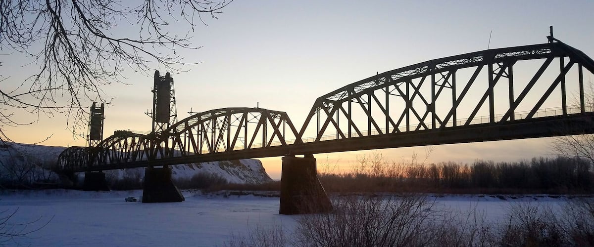 A steel bridge over a snow-covered river with bare trees and a distant hill.