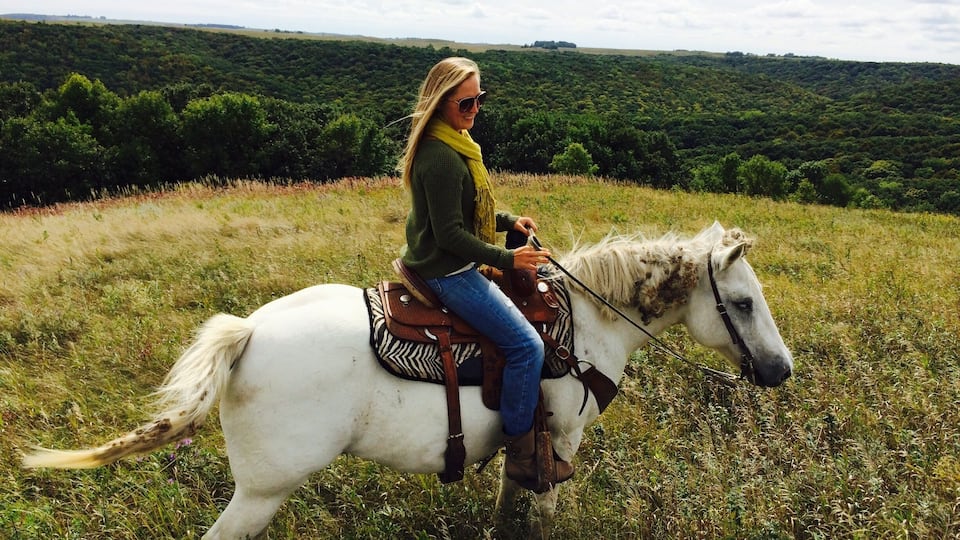 A person riding a white horse in a grassy field with a forested hill in the background.