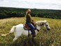A person riding a white horse in a grassy field with a forested hill in the background.