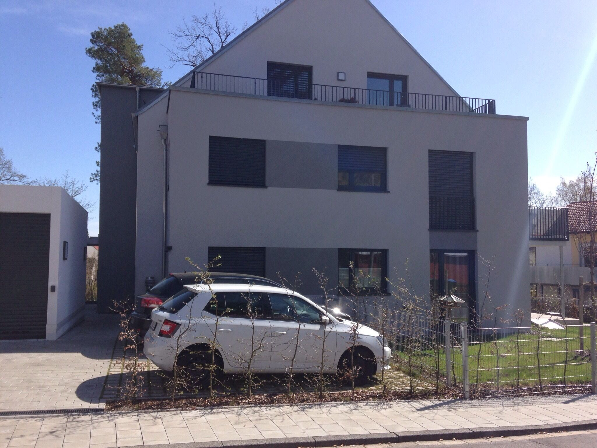 A modern two-story residential building with a white facade and dark shutters, a parked white car, and a fenced yard with sparse vegetation.