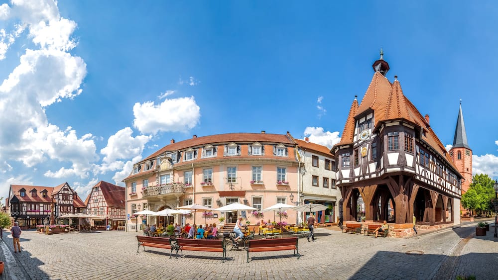 Ein Kopfsteinpflasterplatz mit traditionellen Fachwerkhäusern, einem Kirchturm und Außenbereichs-Sitzgelegenheiten.