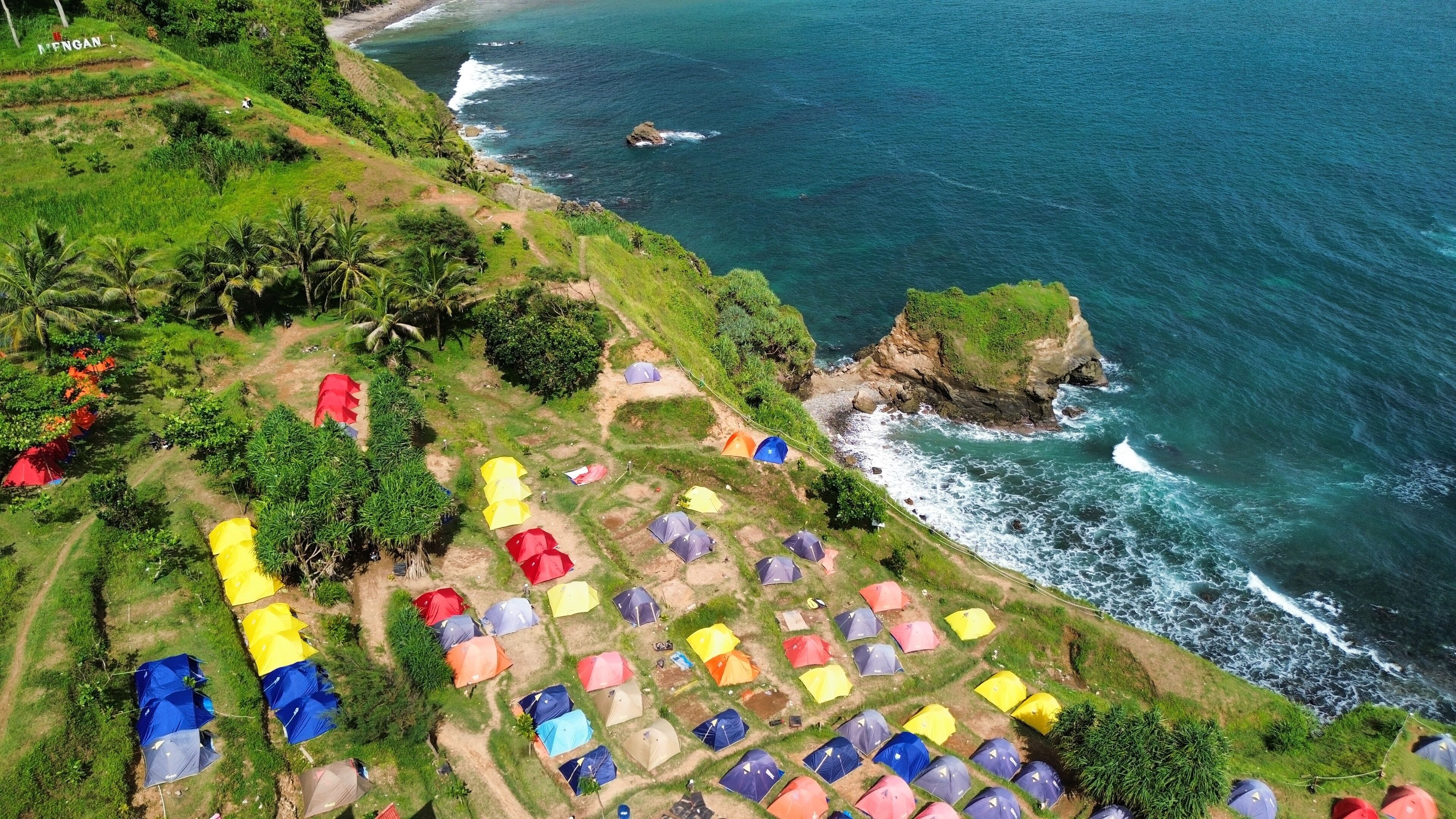 An aerial view of a coastal area with numerous colorful tents set up on grassy terrain, a rocky outcrop, and the ocean waves crashing against the shore.