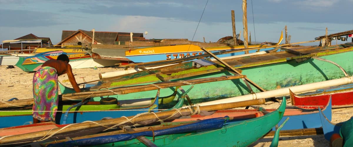 A beach with several colorful boats, a person in a colorful swimsuit, and a few buildings in the background.
