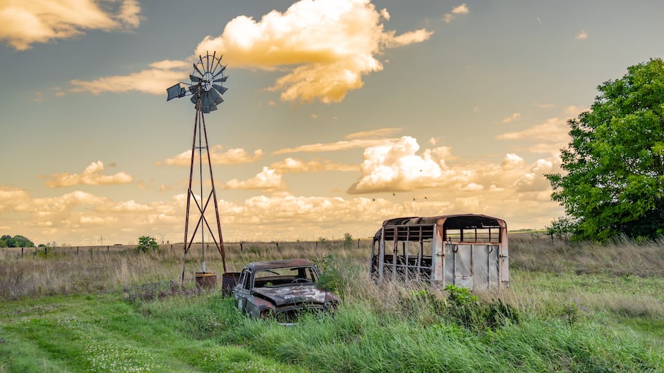 A windmill, an old car, and a wooden structure in a grassy field with a tree and a partly cloudy sky.