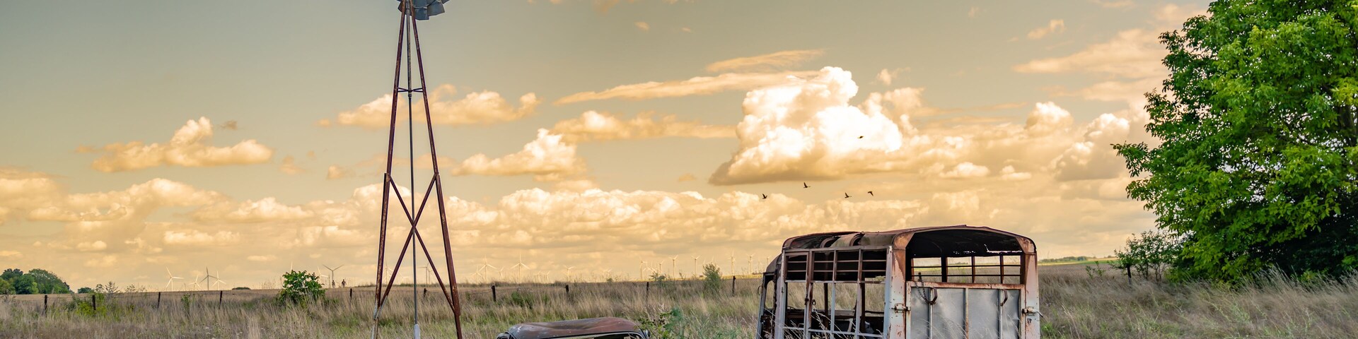 A windmill, an old car, and a wooden structure in a grassy field with a tree and a partly cloudy sky.