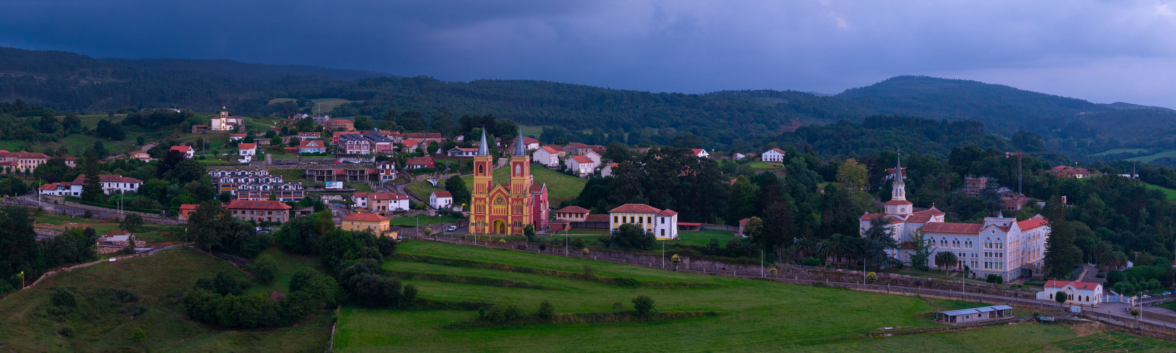 A town with a prominent church, several residential buildings, and rolling hills in the background.