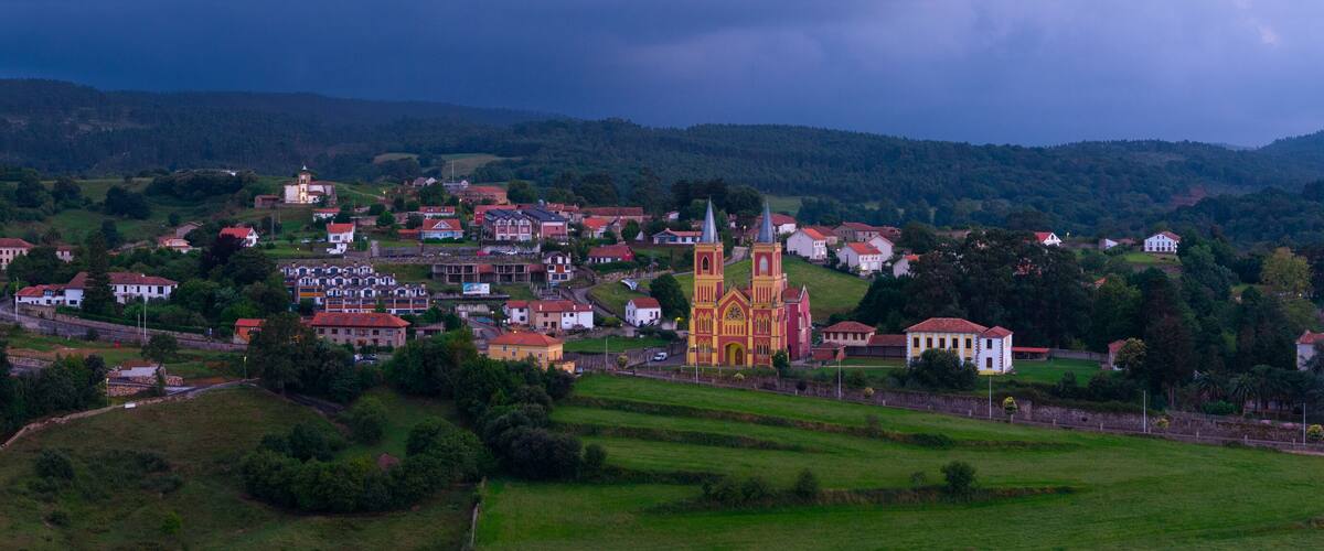 A town with a prominent church, several residential buildings, and rolling hills in the background.
