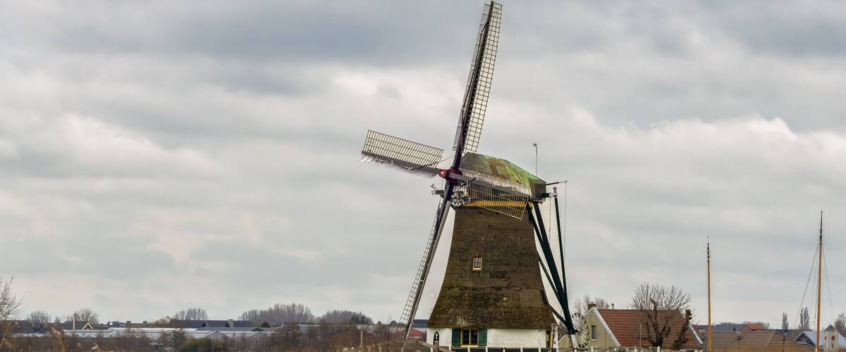 A traditional windmill with a sloped roof, situated near a body of water and surrounded by a rural landscape with houses and fields.