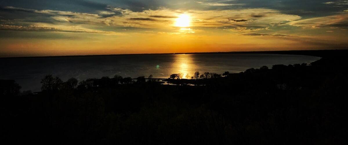 A sunset over a body of water with silhouettes of trees and a distant shoreline.