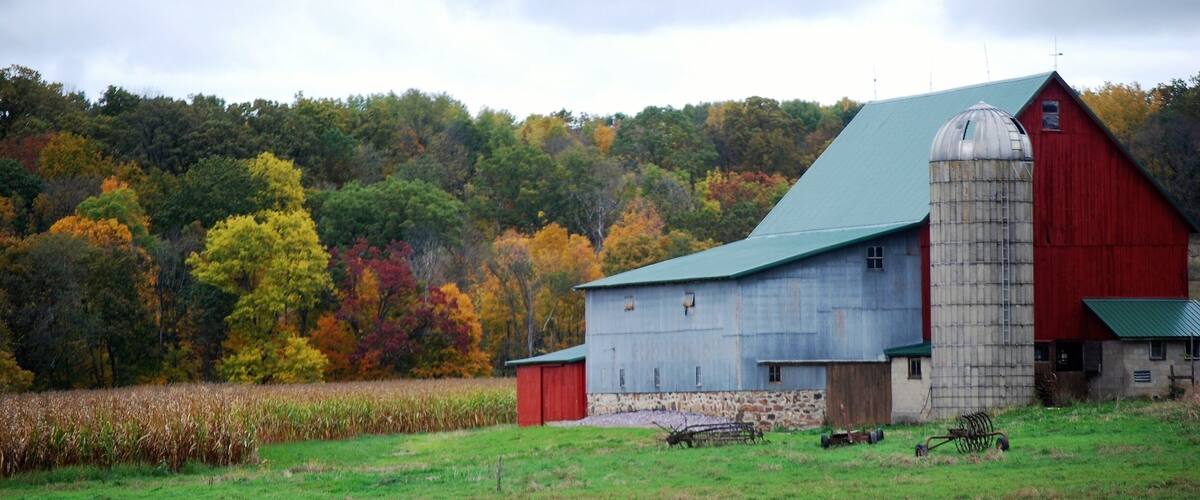 A red barn with a green roof and a silo, surrounded by a field and autumn-colored trees.